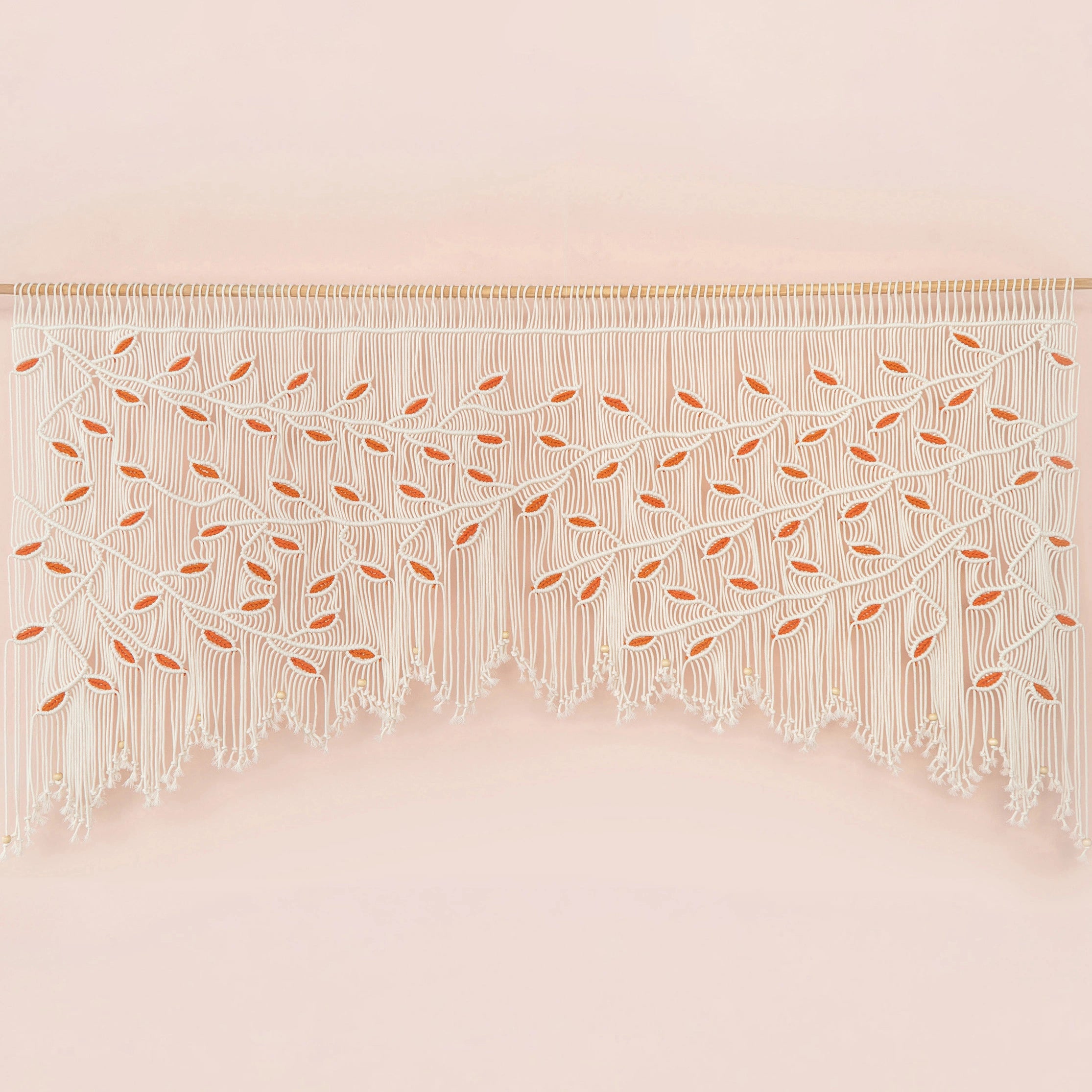 Handcrafted natural beige macrame wall hanging featuring an intricate botanical canopy of cream cords and vibrant orange leaves, hung above a rust-colored velvet sofa.